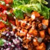 Turkey Taco Sweet Potato Bowl with seasoned ground turkey, roasted sweet potato cubes, black beans, red onion, cherry tomatoes, leafy greens, cilantro, shredded cheese, and creamy lime sauce in a black bowl.