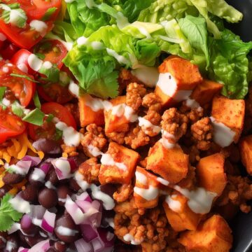 Turkey Taco Sweet Potato Bowl with seasoned ground turkey, roasted sweet potato cubes, black beans, red onion, cherry tomatoes, leafy greens, cilantro, shredded cheese, and creamy lime sauce in a black bowl.