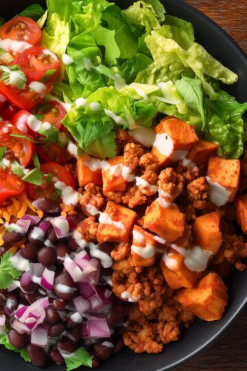 Turkey Taco Sweet Potato Bowl with seasoned ground turkey, roasted sweet potato cubes, black beans, red onion, cherry tomatoes, leafy greens, cilantro, shredded cheese, and creamy lime sauce in a black bowl.