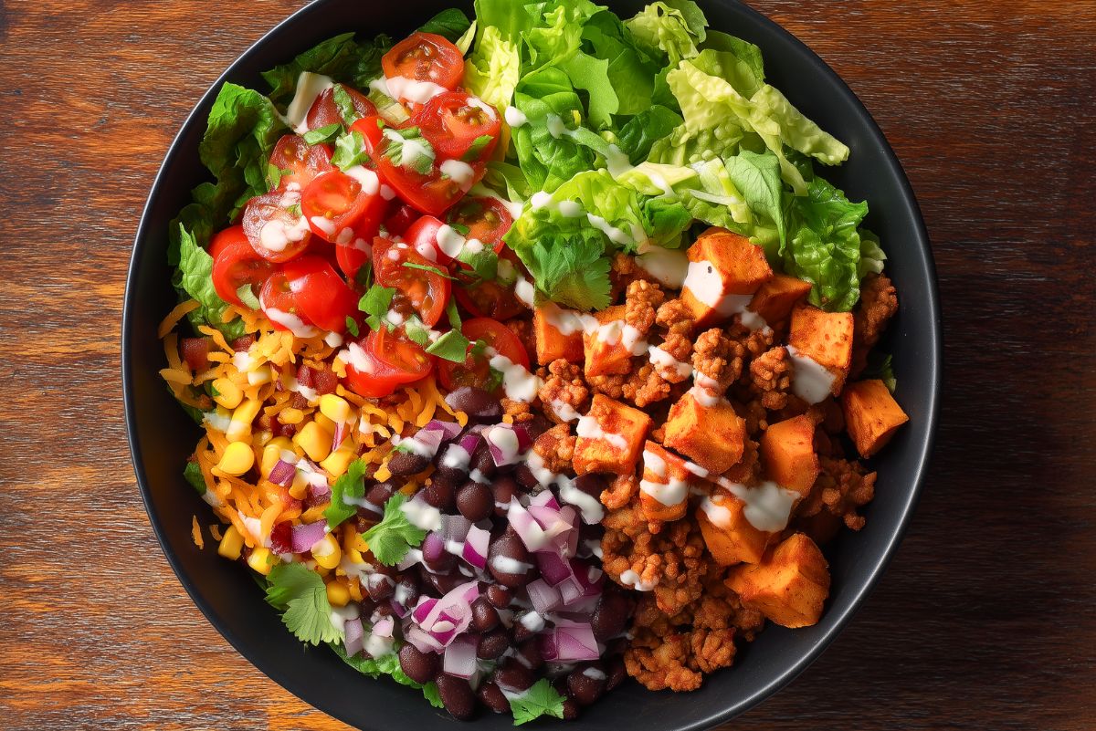 High protein Turkey Taco Sweet Potato Bowl with seasoned ground turkey, roasted sweet potatoes, black beans, corn, red onion, cherry tomatoes, leafy greens, shredded cheese, cilantro, and creamy lime sauce in a black bowl.