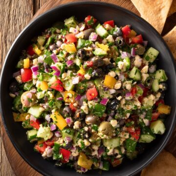 Overhead view of veggie grinder chopped salad in a black bowl with cucumbers, colorful bell peppers, red onion, olives, herbs, and crumbled cheese, surrounded by garlic pita chips on a rustic wooden table.