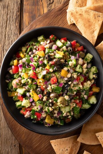 Overhead view of veggie grinder chopped salad in a black bowl with cucumbers, colorful bell peppers, red onion, olives, herbs, and crumbled cheese, surrounded by garlic pita chips on a rustic wooden table.