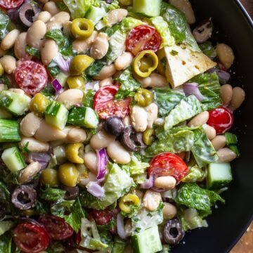 White bean grinder chopped salad with crisp romaine lettuce, cucumber, cherry tomatoes, green and black olives, red onion, fresh herbs, and toasted pita chips served in a black bowl.