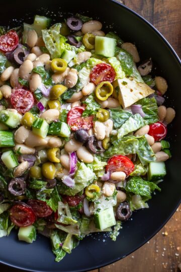 White bean grinder chopped salad with crisp romaine lettuce, cucumber, cherry tomatoes, green and black olives, red onion, fresh herbs, and toasted pita chips served in a black bowl.