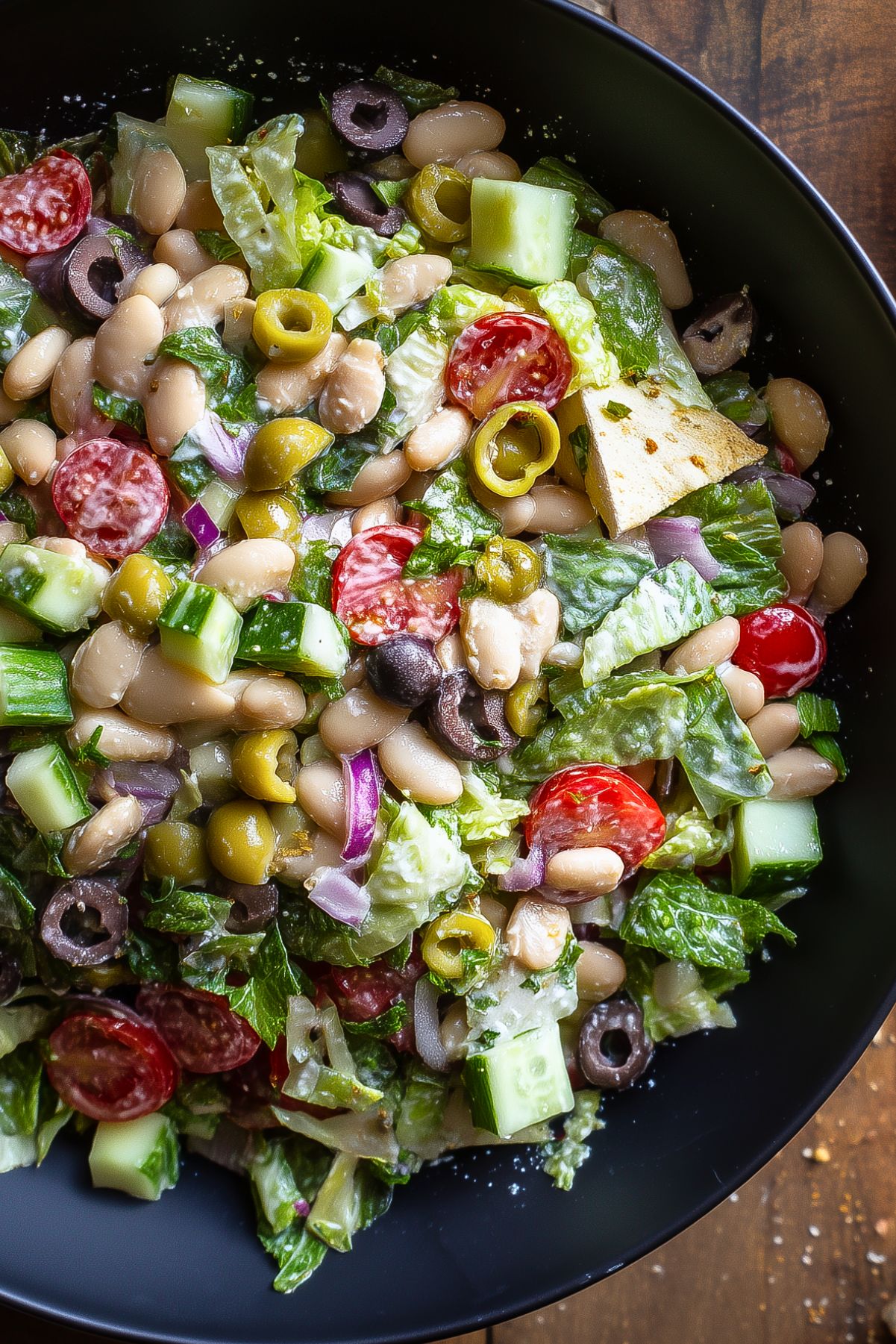 White bean grinder chopped salad with crisp romaine lettuce, cucumber, cherry tomatoes, green and black olives, red onion, fresh herbs, and toasted pita chips served in a black bowl.