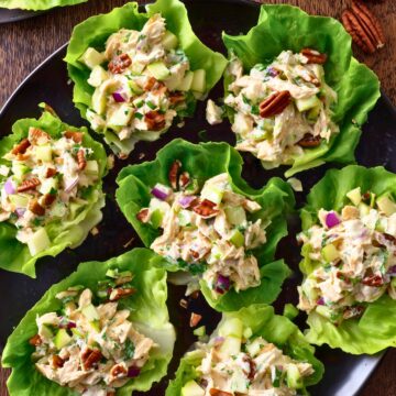 Overhead view of apple cheddar chicken lettuce wraps arranged on a black platter, topped with creamy chicken salad, diced apples, celery, red onion, and pecans, with whole and sliced apples nearby.
