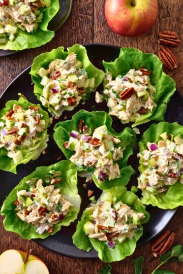 Overhead view of apple cheddar chicken lettuce wraps arranged on a black platter, topped with creamy chicken salad, diced apples, celery, red onion, and pecans, with whole and sliced apples nearby.