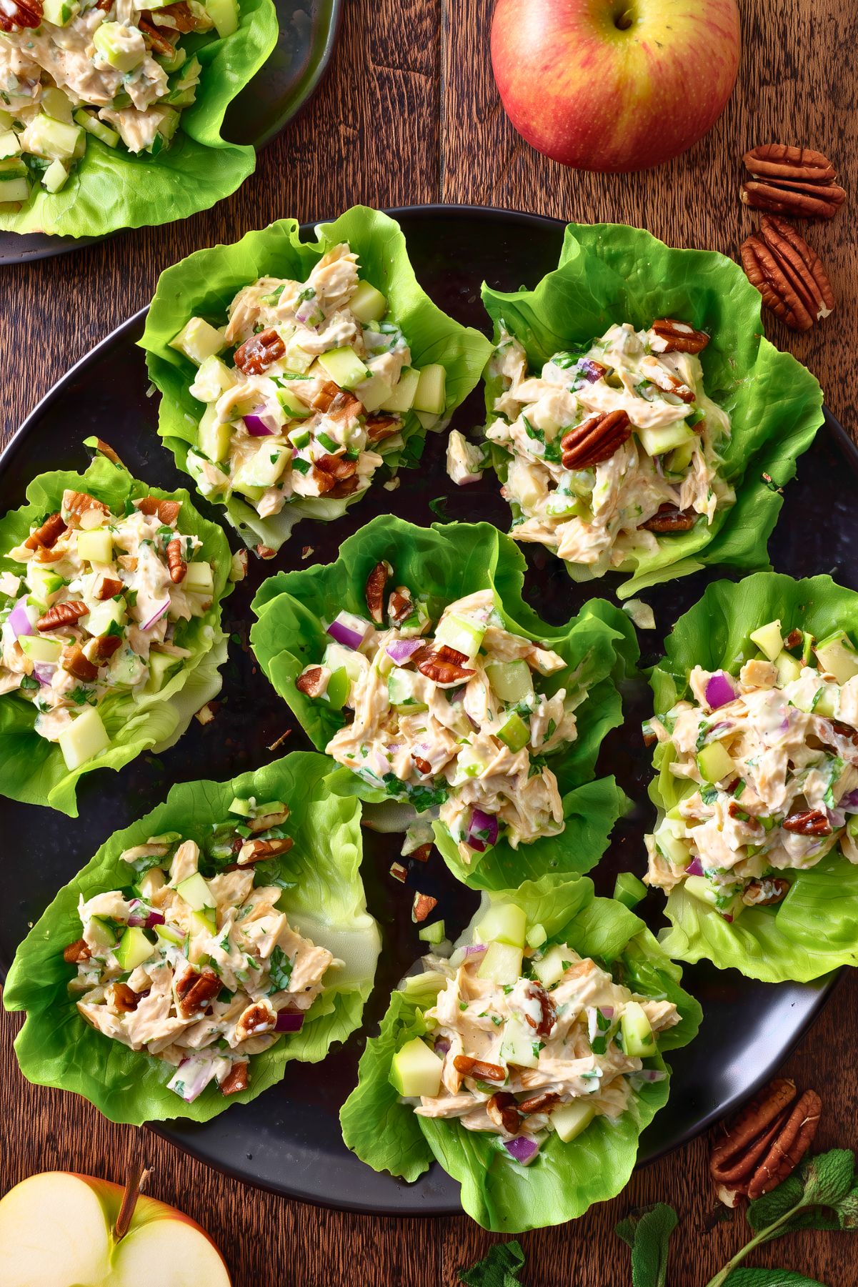 Overhead view of apple cheddar chicken lettuce wraps arranged on a black platter, topped with creamy chicken salad, diced apples, celery, red onion, and pecans, with whole and sliced apples nearby.