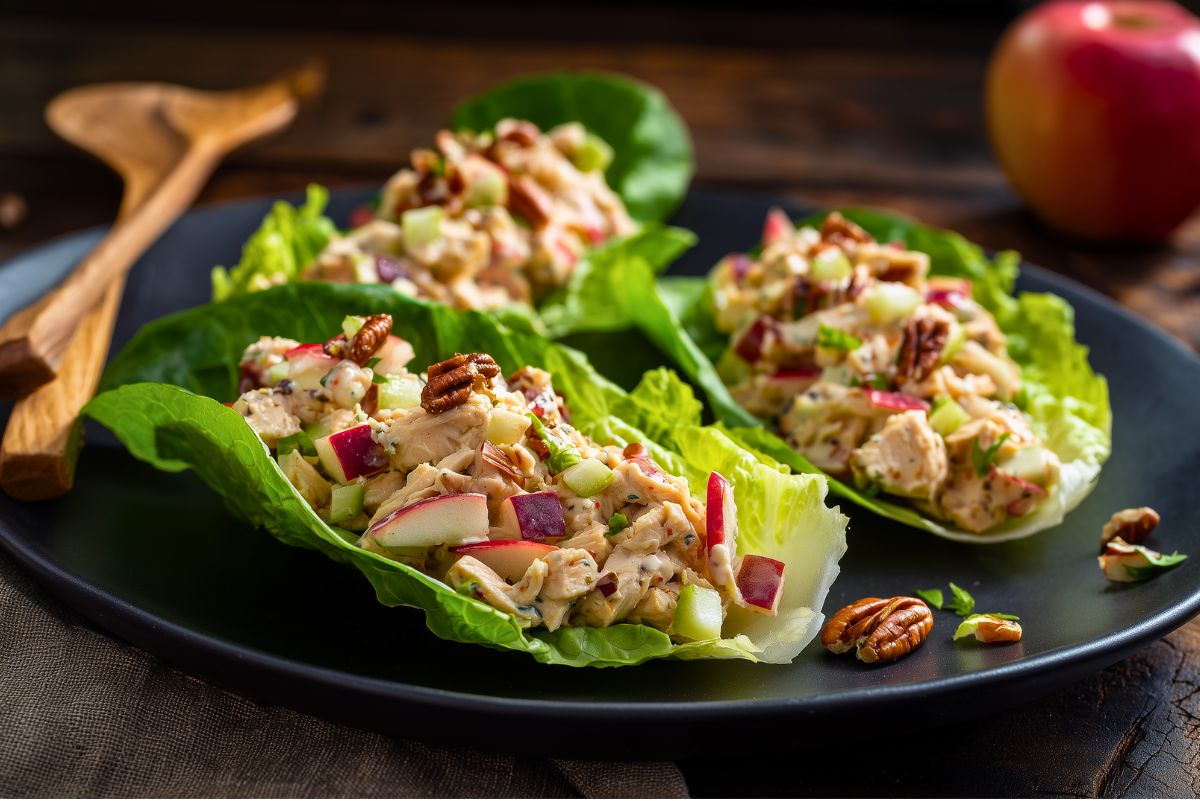Apple cheddar chicken lettuce wraps on a black plate, filled with creamy chicken salad, diced red apple, celery, cheddar cheese, and pecans. A whole red apple and wooden salad servers sit in the background.