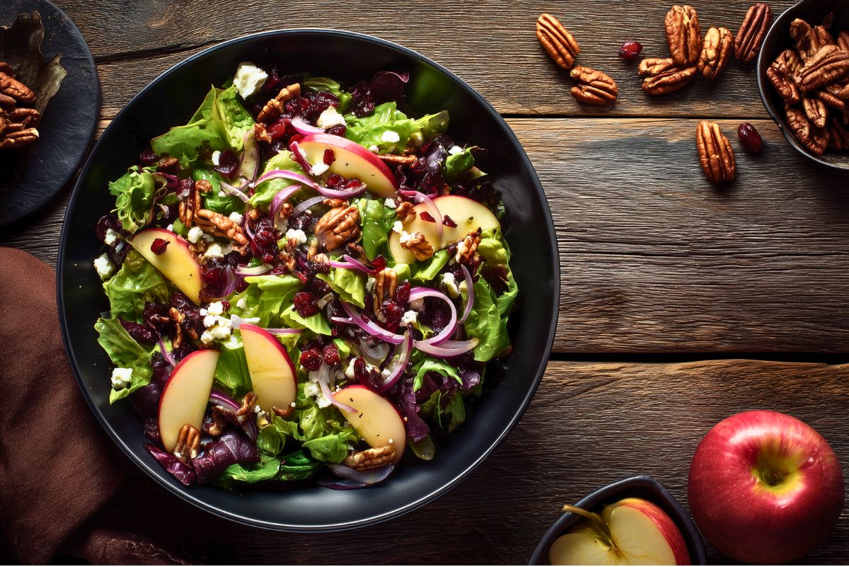 Apple Cranberry Pecan Salad served in a black bowl with mixed greens, sliced red apples, dried cranberries, pecans, crumbled feta, and thin red onion on a dark rustic wood table.