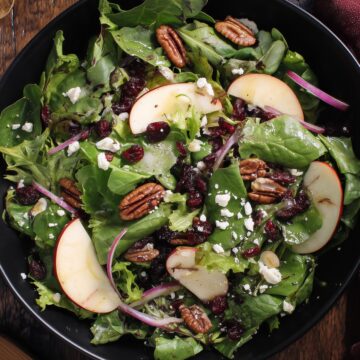 Overhead view of Apple Cranberry Pecan Salad with leafy greens, apple slices, dried cranberries, pecans, feta cheese, and red onion in a black bowl on a rustic wooden table.