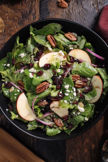 Overhead view of Apple Cranberry Pecan Salad with leafy greens, apple slices, dried cranberries, pecans, feta cheese, and red onion in a black bowl on a rustic wooden table.