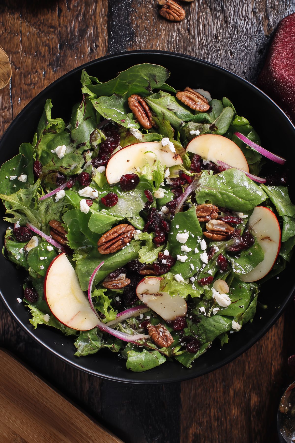 Overhead view of Apple Cranberry Pecan Salad with leafy greens, apple slices, dried cranberries, pecans, feta cheese, and red onion in a black bowl on a rustic wooden table.