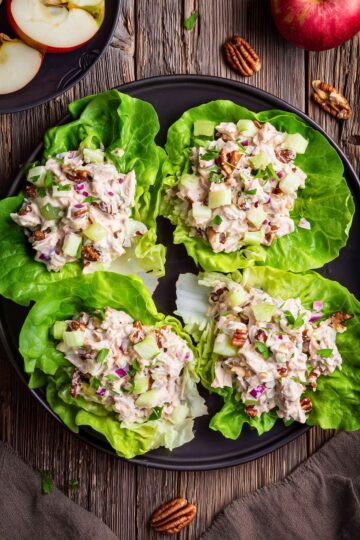 Overhead view of four apple pecan chicken lettuce wraps on a black plate, filled with creamy chicken salad, diced apple, celery, red onion, pecans, and herbs. Fresh apples and pecans sit on a rustic wood table.