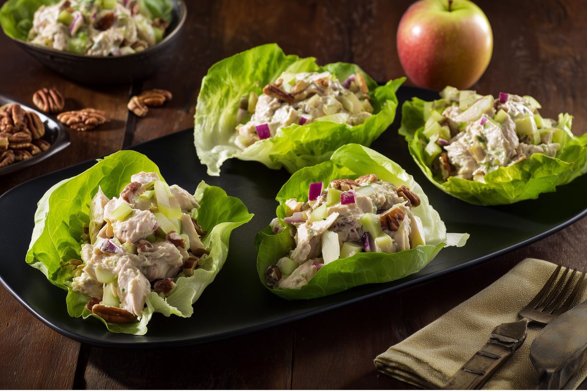 Four apple pecan chicken lettuce wraps filled with creamy chicken salad, diced green apple, red onion, celery, and pecans on a black serving platter. A red apple, extra pecans, and a bowl of chicken salad sit on a dark rustic table.