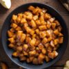 Overhead view of homemade apple pie filling with tender diced apples coated in a glossy cinnamon sauce in a black bowl, surrounded by sliced apples on a rustic wood table.