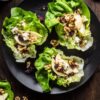 Three apple walnut and brie lettuce wraps on a black plate with crisp butter lettuce, sliced apples, walnuts, dried cranberries, creamy brie, and dressing on a rustic wood table.