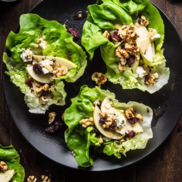 Three apple walnut and brie lettuce wraps on a black plate with crisp butter lettuce, sliced apples, walnuts, dried cranberries, creamy brie, and dressing on a rustic wood table.