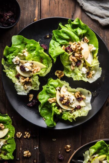 Three apple walnut and brie lettuce wraps on a black plate with crisp butter lettuce, sliced apples, walnuts, dried cranberries, creamy brie, and dressing on a rustic wood table.