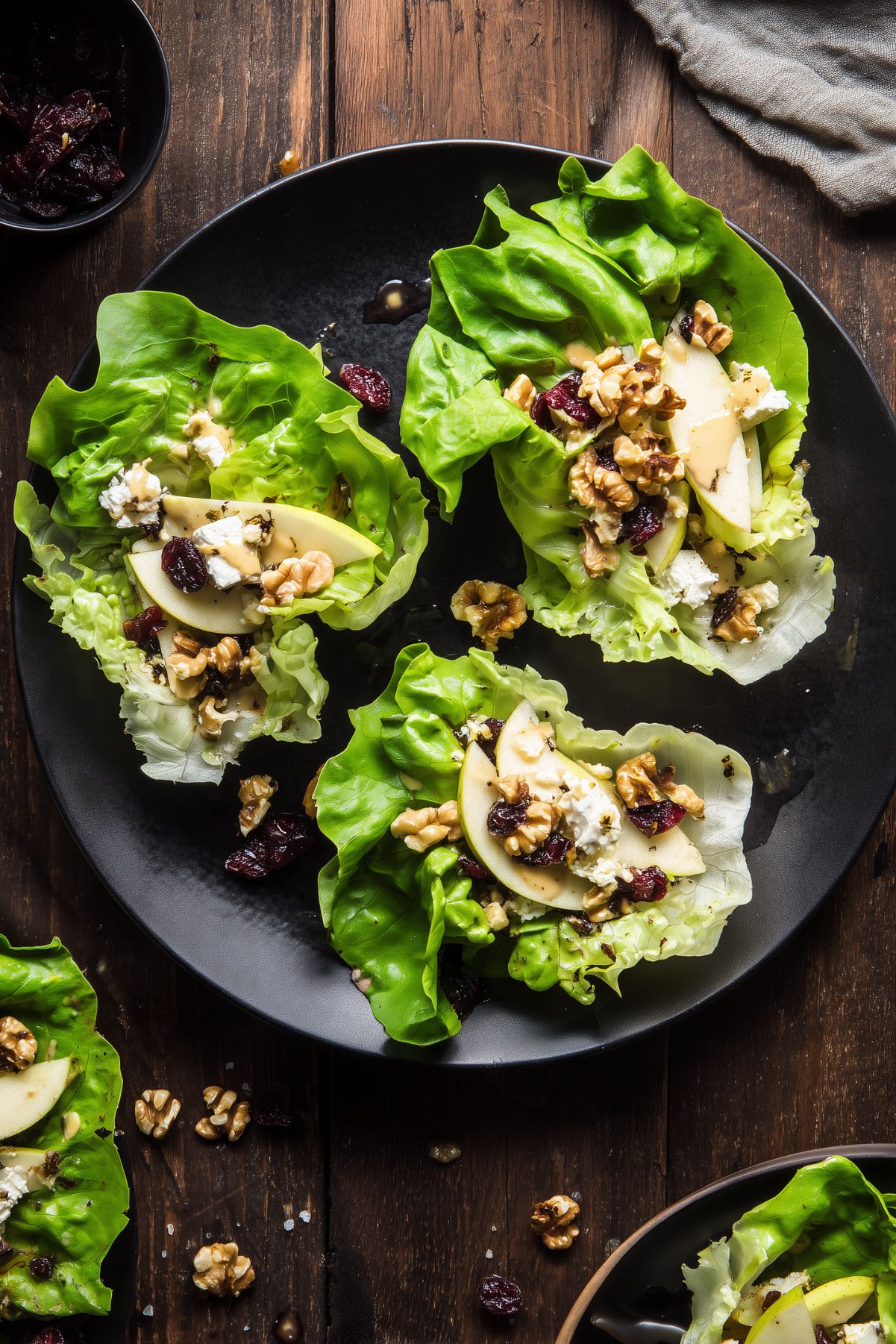 Three apple walnut and brie lettuce wraps on a black plate with crisp butter lettuce, sliced apples, walnuts, dried cranberries, creamy brie, and dressing on a rustic wood table.