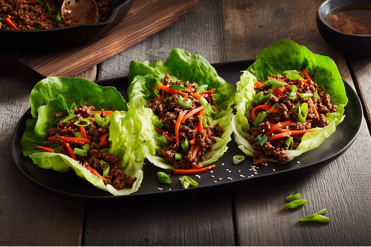 Three Asian Ground Beef Lettuce Wraps filled with seasoned beef, carrots, red pepper, green onions, and sesame seeds, served on a black platter on a rustic wood table.