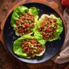 Overhead view of three Asian Ground Beef Lettuce Wraps filled with savory beef, carrots, red pepper, green onions, and sesame seeds on a black plate over a rustic wood table.