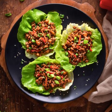 Overhead view of three Asian Ground Beef Lettuce Wraps filled with savory beef, carrots, red pepper, green onions, and sesame seeds on a black plate over a rustic wood table.