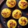Overhead view of bacon cheeseburger egg bites topped with crumbled beef, bacon, cheddar, and herbs on a black plate over a rustic wooden table.