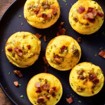 Overhead view of bacon cheeseburger egg bites topped with crumbled beef, bacon, cheddar, and herbs on a black plate over a rustic wooden table.