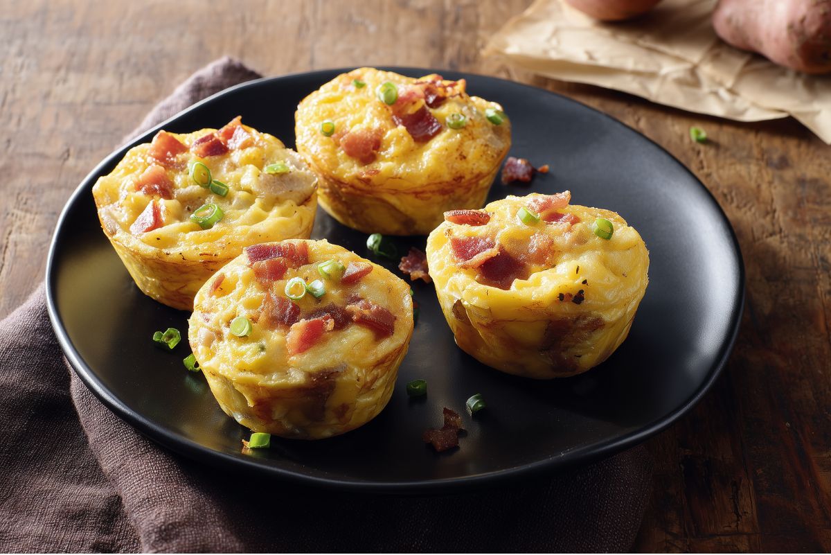 Four bacon and potato egg bites with melted cheddar and green onions served on a black plate over a rustic wooden table.
