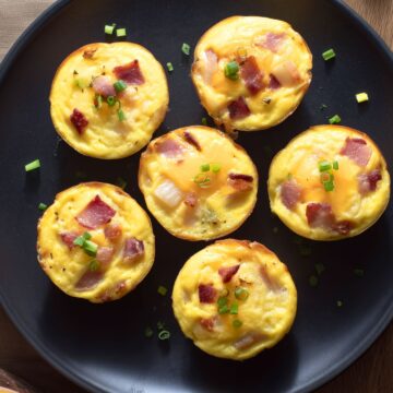 Six bacon and potato egg bites with cheddar cheese and green onions arranged on a black plate, photographed from overhead.