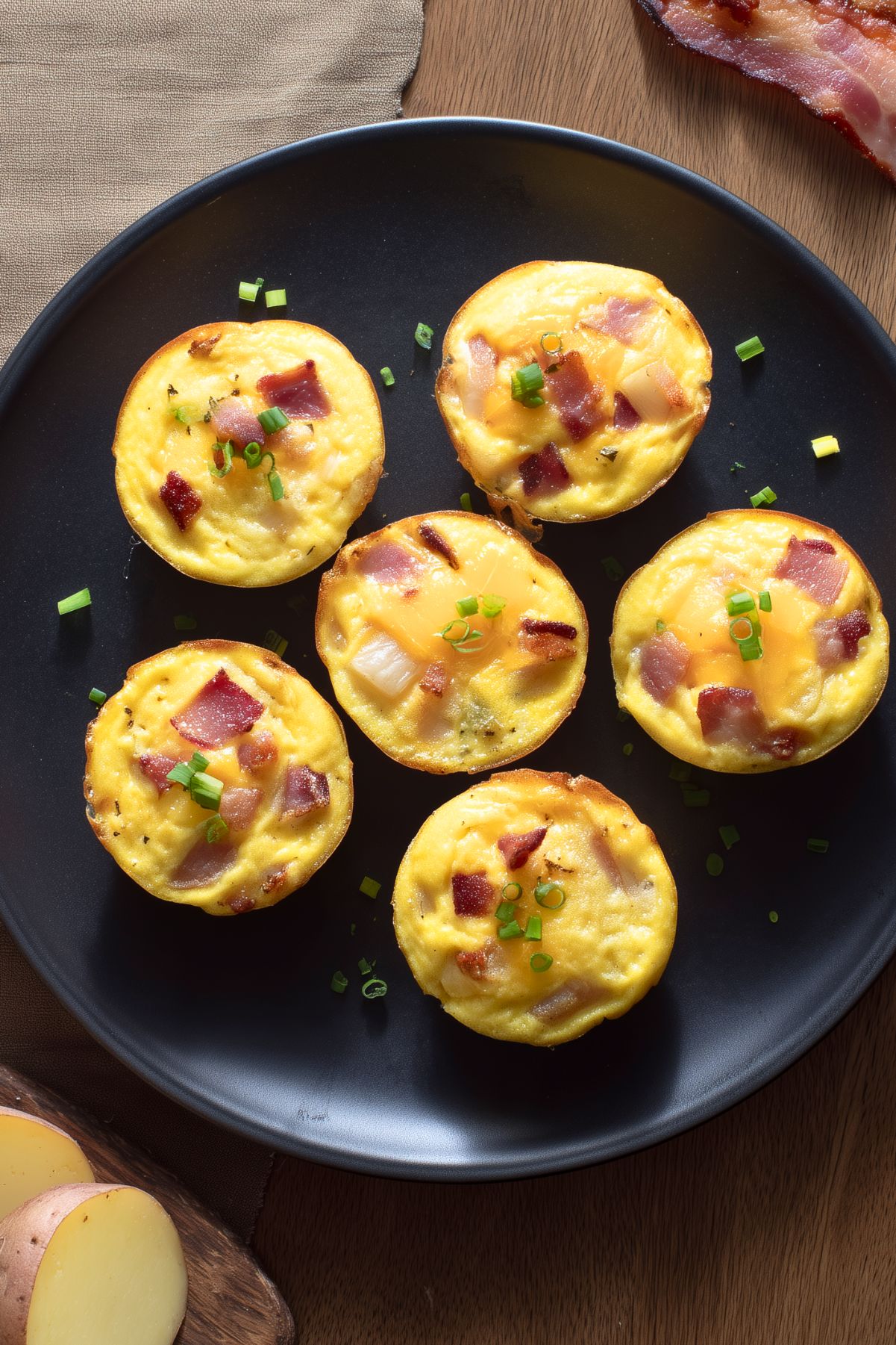 Six bacon and potato egg bites with cheddar cheese and green onions arranged on a black plate, photographed from overhead.