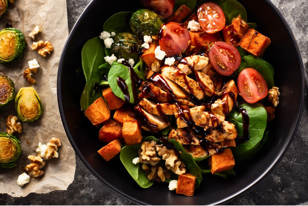 Balsamic Chicken Sweet Potato Bowl in a black bowl with sliced chicken, roasted sweet potatoes, Brussels sprouts, spinach, cherry tomatoes, walnuts, crumbled cheese, and balsamic glaze.