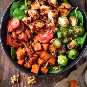 Overhead Balsamic Chicken Sweet Potato Bowl with glazed chicken, roasted sweet potatoes, Brussels sprouts, spinach, tomatoes, and walnuts in a black bowl on rustic wood.