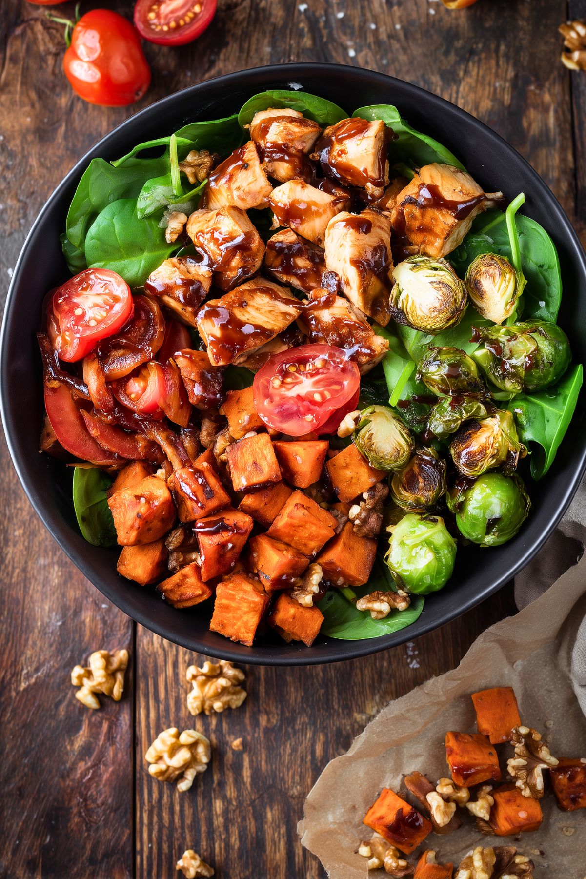 Overhead Balsamic Chicken Sweet Potato Bowl with glazed chicken, roasted sweet potatoes, Brussels sprouts, spinach, tomatoes, and walnuts in a black bowl on rustic wood.