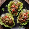 Three beef taco lettuce wraps with seasoned ground beef, diced tomatoes, red onion, shredded cheese, and cilantro arranged on a black plate over a rustic wood table.