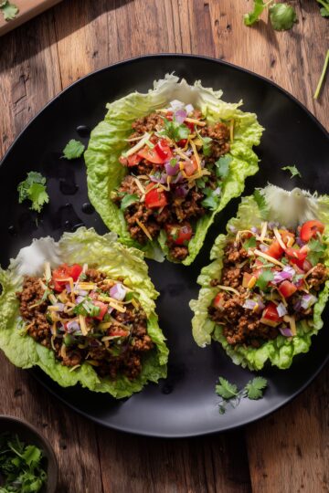 Three beef taco lettuce wraps with seasoned ground beef, diced tomatoes, red onion, shredded cheese, and cilantro arranged on a black plate over a rustic wood table.
