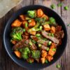 Beef and broccoli sweet potato bowl with tender sliced beef, roasted sweet potato cubes, broccoli, green onions, and sesame seeds in a savory sauce, served in a black bowl.