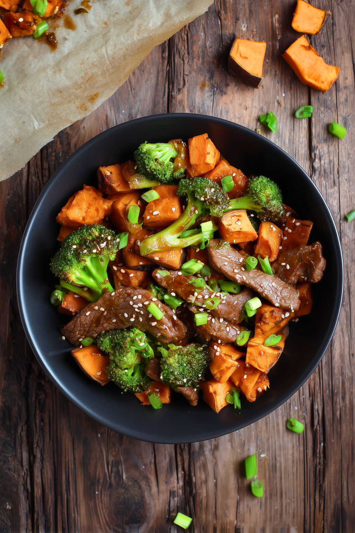 Beef and broccoli sweet potato bowl with tender sliced beef, roasted sweet potato cubes, broccoli, green onions, and sesame seeds in a savory sauce, served in a black bowl.