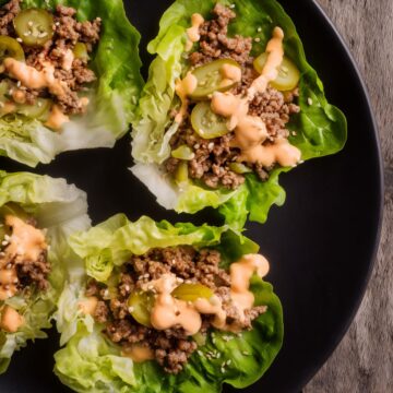 Overhead view of Big Mac lettuce wraps filled with ground beef and sliced pickles, drizzled with creamy burger sauce on a black plate over a rustic wood table.