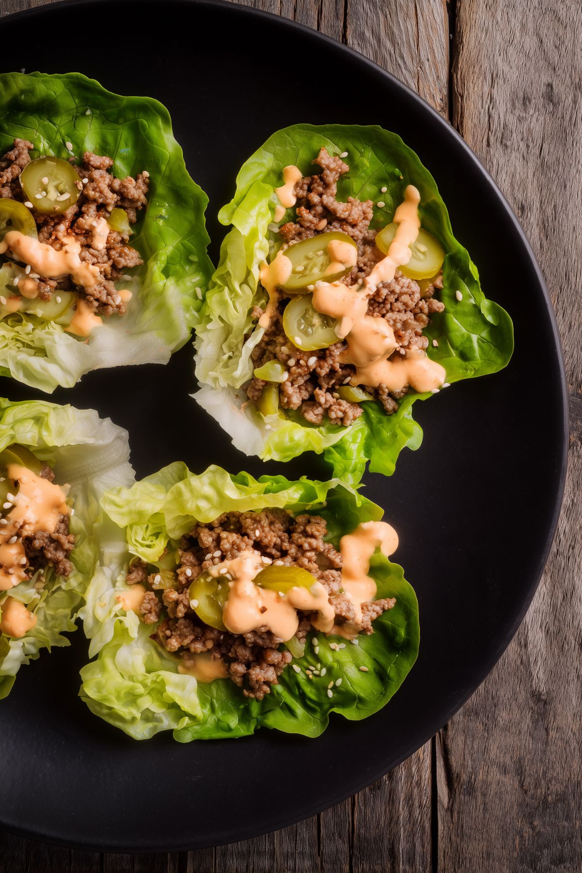 Overhead view of Big Mac lettuce wraps filled with ground beef and sliced pickles, drizzled with creamy burger sauce on a black plate over a rustic wood table.