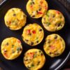 Overhead view of seven black bean and corn egg bites with red bell pepper, green onion, and melted cheese arranged on a black plate over a rustic wood table.