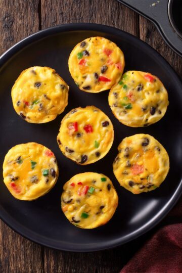 Overhead view of seven black bean and corn egg bites with red bell pepper, green onion, and melted cheese arranged on a black plate over a rustic wood table.
