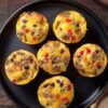 Overhead view of six Breakfast Burrito Egg Bites with sausage, black beans, peppers, green chiles, and melted cheese arranged on a black plate over a dark rustic wood table.