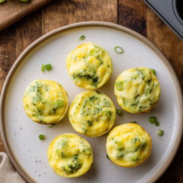 Six broccoli cheddar egg bites arranged on a white plate over rustic wood, with fresh broccoli on a cutting board and a muffin pan nearby.