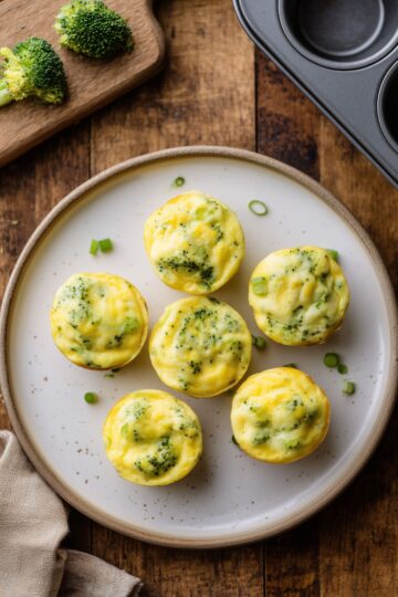 Six broccoli cheddar egg bites arranged on a white plate over rustic wood, with fresh broccoli on a cutting board and a muffin pan nearby.