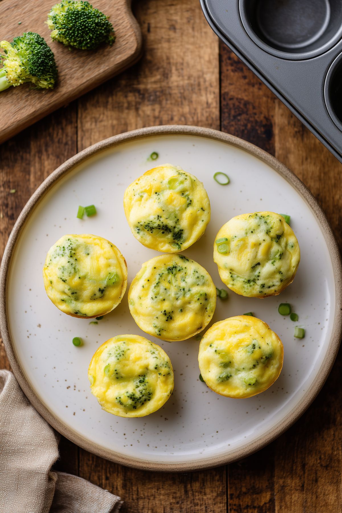 Six broccoli cheddar egg bites arranged on a white plate over rustic wood, with fresh broccoli on a cutting board and a muffin pan nearby.