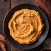Overhead view of brown butter mashed sweet potatoes in a black bowl on a dark rustic wood table, accented with autumn leaves, a brown napkin, and a serving spoon.