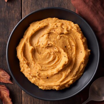 Overhead view of brown butter mashed sweet potatoes in a black bowl on a dark rustic wood table, accented with autumn leaves, a brown napkin, and a serving spoon.