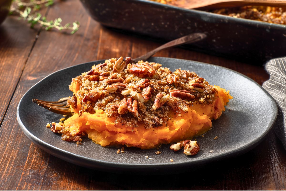A serving of creamy butternut squash casserole on a dark plate, topped with toasted pecans and a golden breadcrumb mixture.